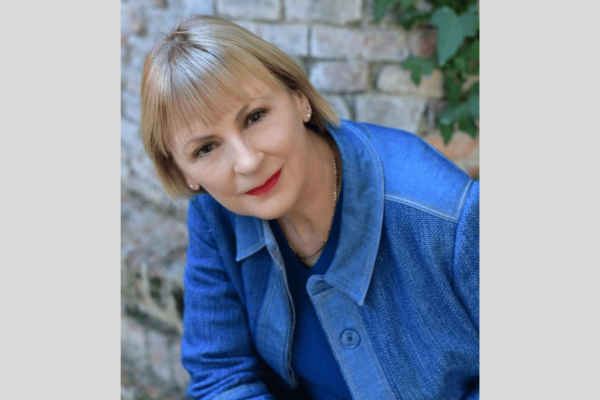 Portrait of Lindsay Nicholson, a white woman with short blonde hair wearing a blue shirt over a blue top sat with a brick wall and some foliage behind her